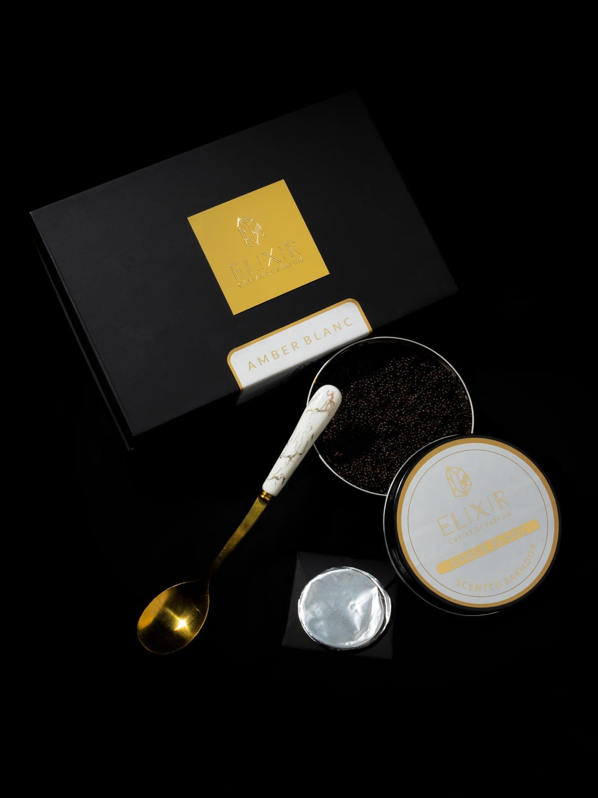 The Amber Blanc box open on black, its white-lidded tin and a spoon of pearls